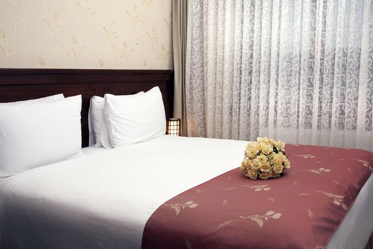 A neatly made bed with white and maroon bedding, a floral-patterned throw, and a bouquet of flowers on the bed.