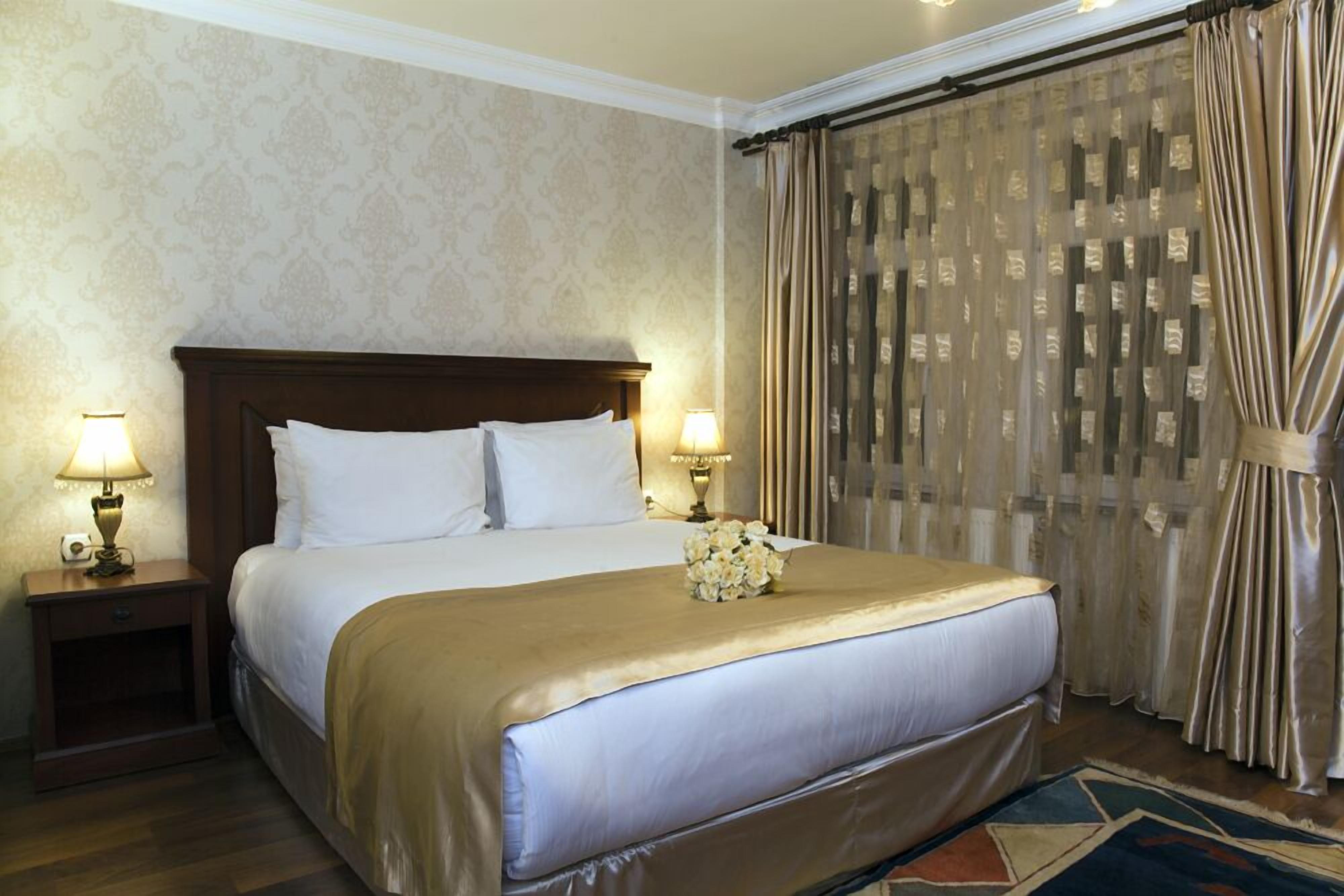 A hotel room with a large bed, two bedside tables with lamps, a decorative vase, and a patterned curtain.