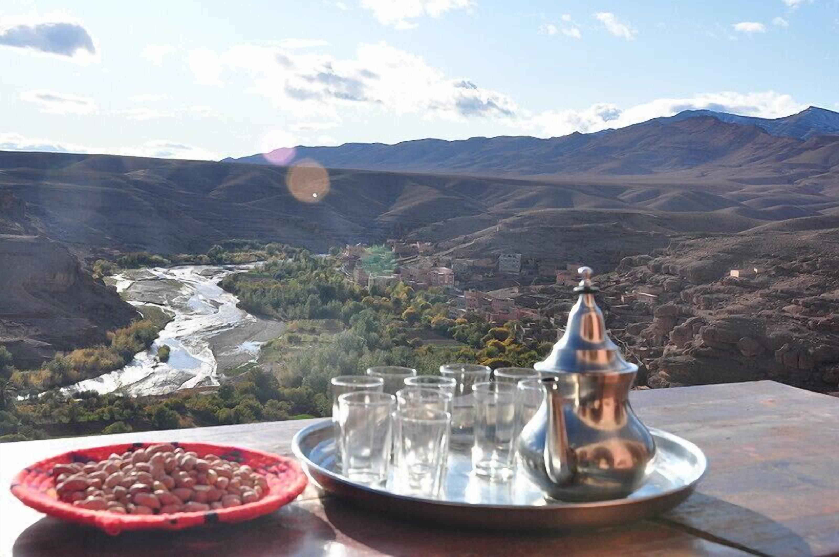 Uma mesa com uma bandeja de copos e uma panela de metal, com uma paisagem montanhosa ao fundo, incluindo um rio e edifícios.
