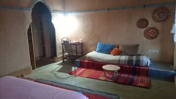 A room with a bed, a chair, a small table, and a sofa. There are decorative plates on the wall and a window with sunlight coming through.