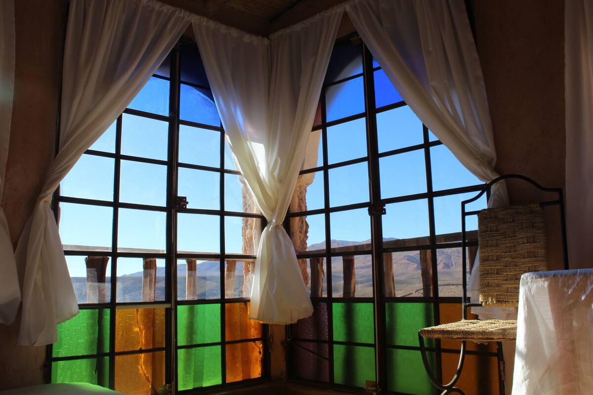 A room with large windows, curtains, a wicker chair, and a small table.