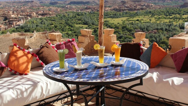 A table with drinks and colorful cushions on a terrace overlooking a scenic landscape.