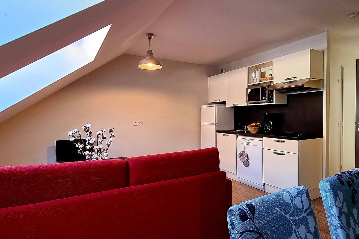 A compact living space with a sloped ceiling, a kitchenette, a dining area with a red sofa and a blue chair, and a small table.