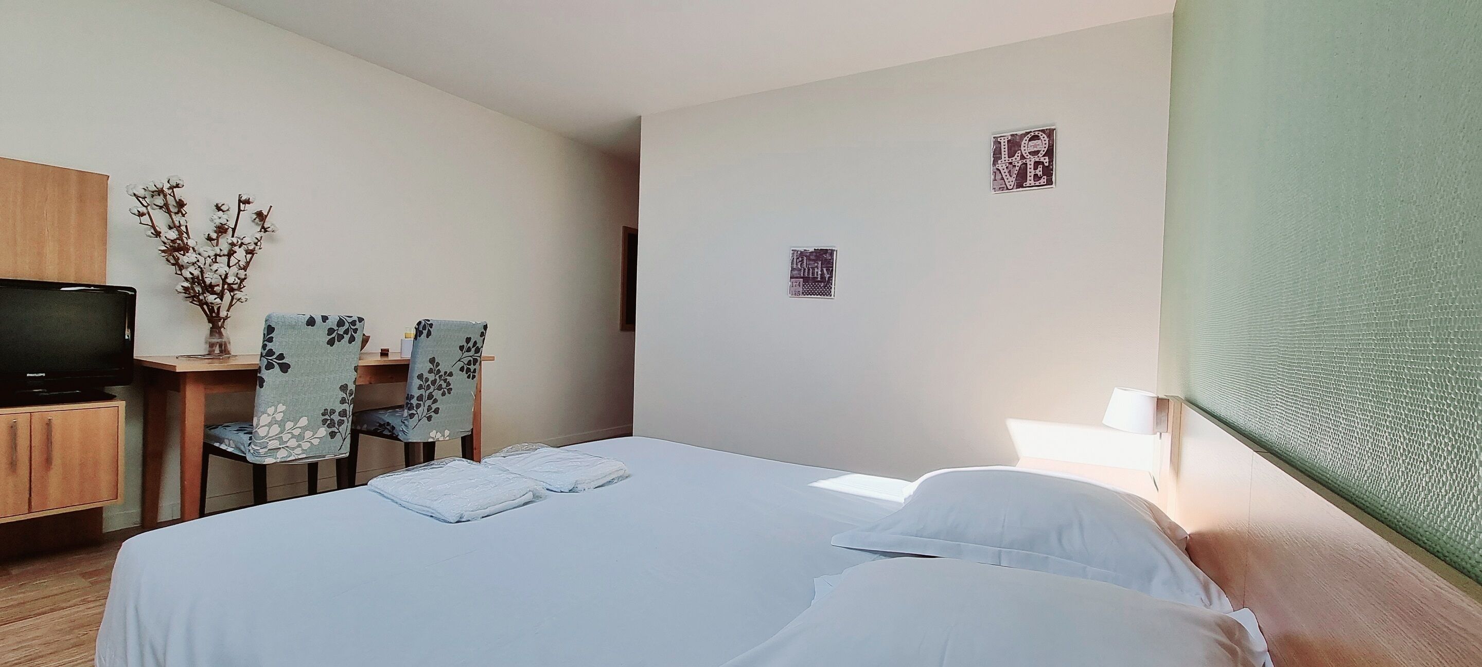 Une chambre d’hôtel avec un lit, deux chaises, un bureau avec une télévision et un vase rempli de fleurs.