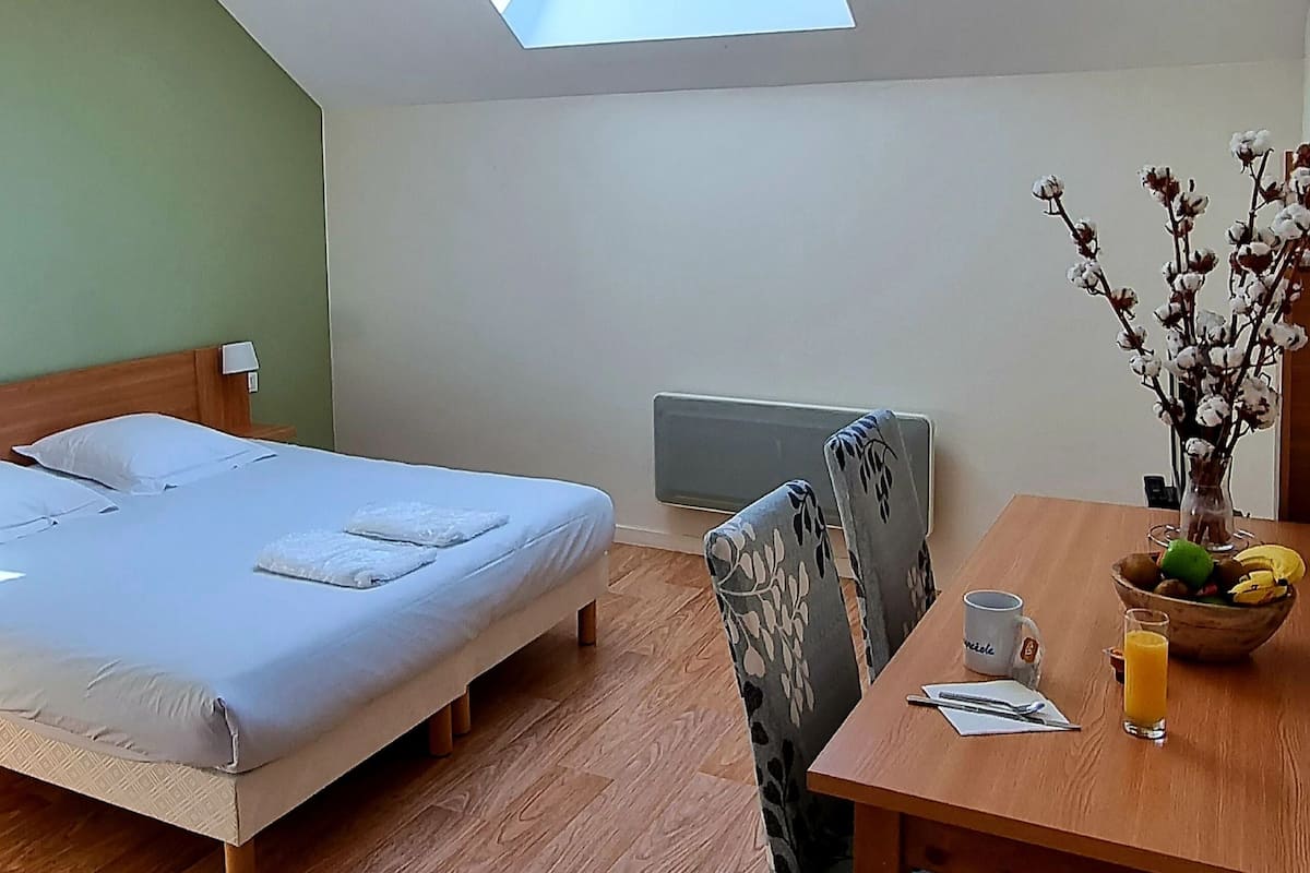 A room with a skylight, a bed, a desk with a chair, a bowl of fruit, and a vase of flowers.