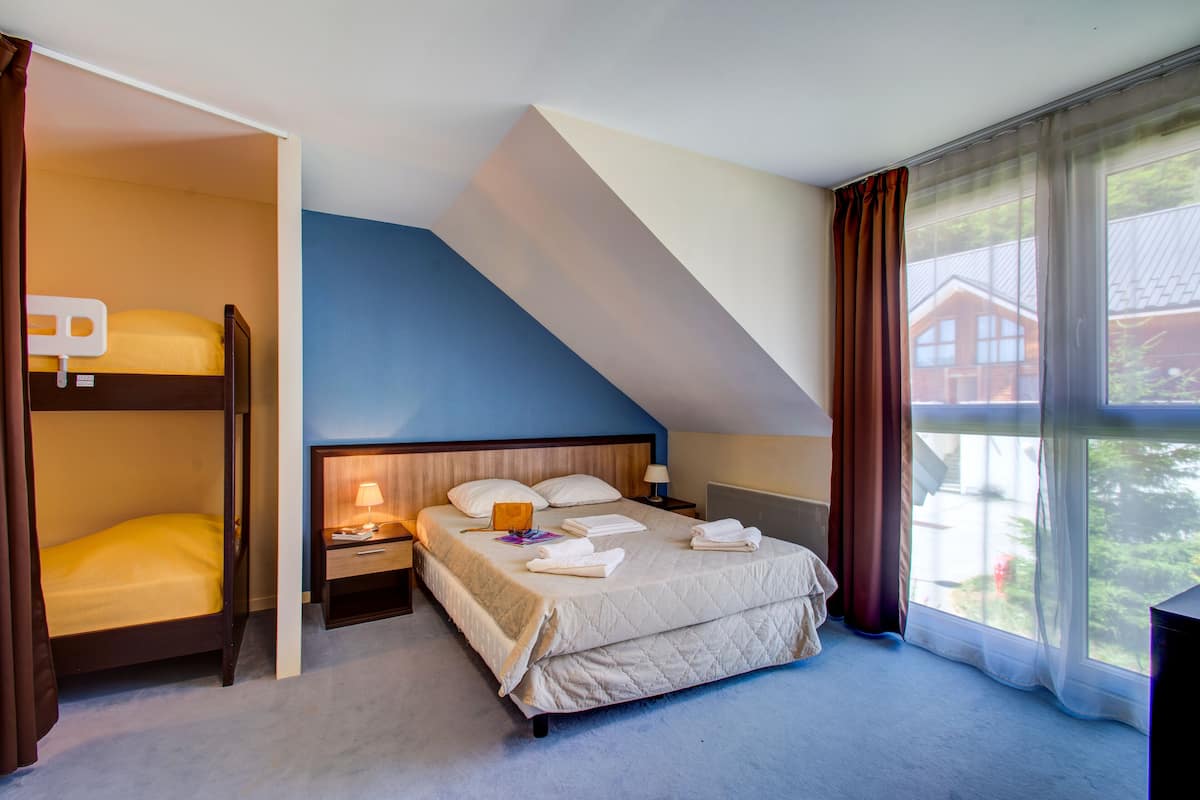 A bedroom with a bunk bed, a single bed, a nightstand, a lamp, and a view of a house through a large window.