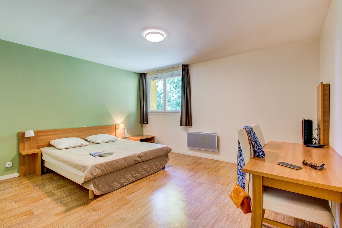 A bedroom with a bed, bedside table, lamp, window, radiator, and a desk with a chair.