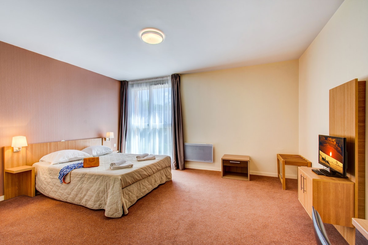 A hotel room with a bed, bedside tables, a TV, a desk, and a window with curtains.