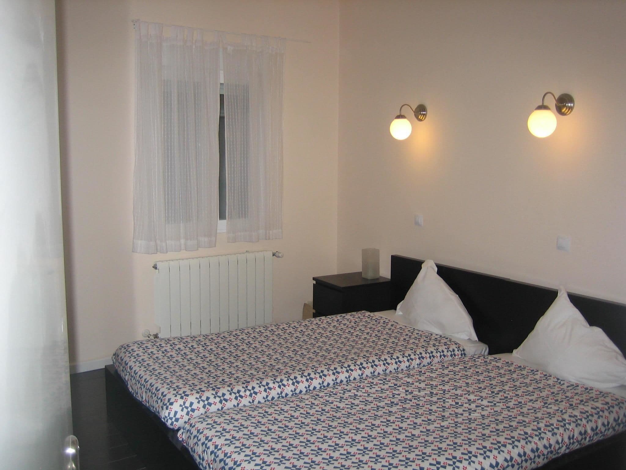 A bedroom with a bed, a window with sheer curtains, a bedside table, and two wall-mounted light fixtures.