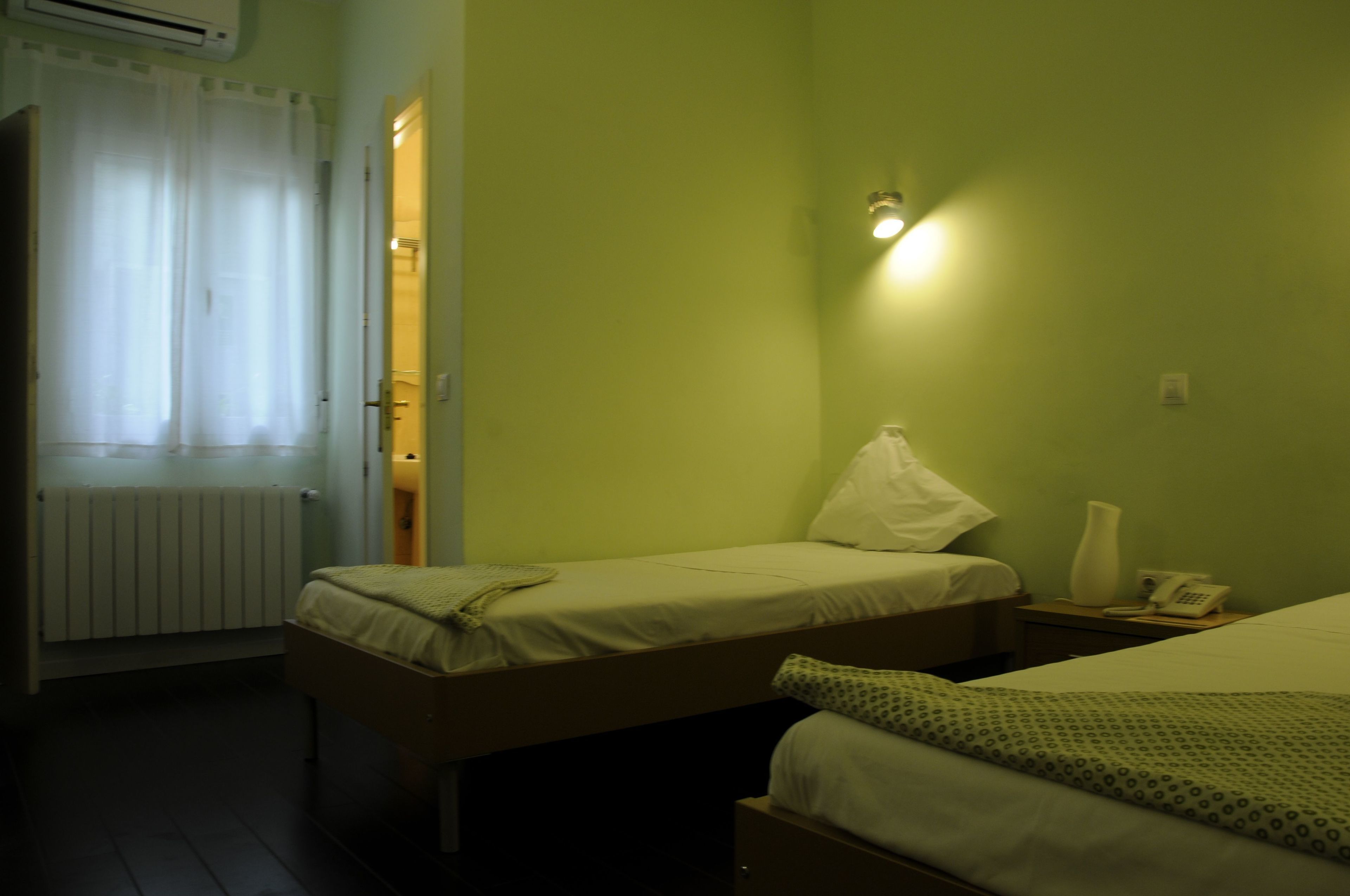 Una habitación de hotel pequeña y sencilla, con dos camas, una mesita de noche con lámpara, un radiador y una ventana con cortinas.