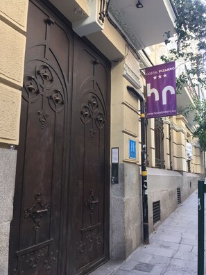 Une grande porte en métal, richement décorée, avec un trou de serrure et une sonnette. Une bannière violette portant l’inscription « HOSTAL PIZARRO » est suspendue au-dessus de la porte.