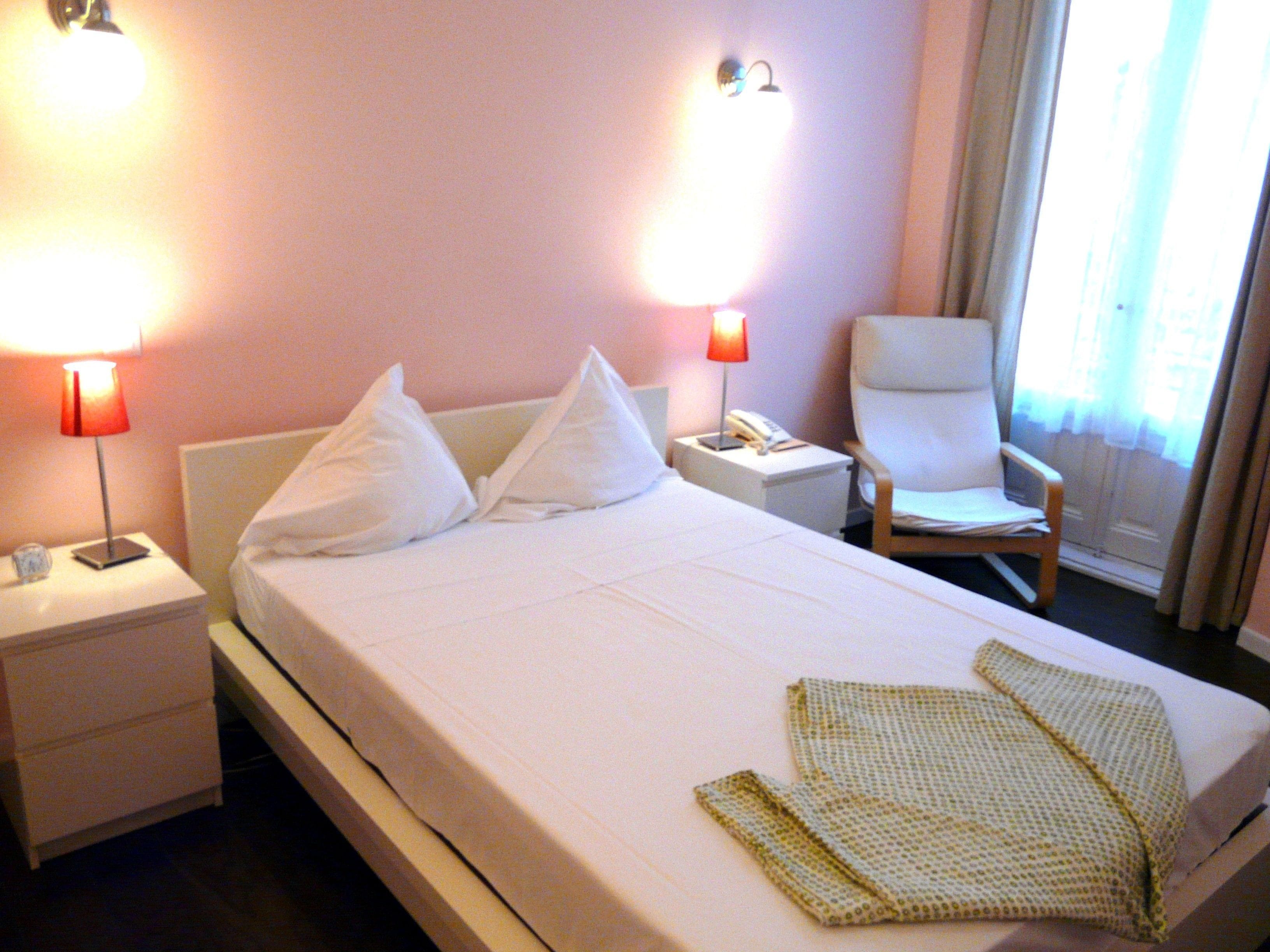 A hotel room with a bed, two bedside lamps, a chair, a nightstand, and a window with curtains.