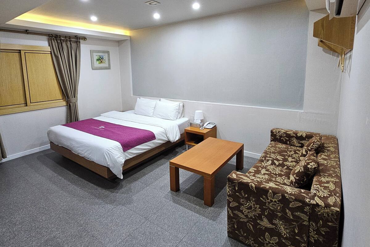 A hotel room with a bed, a small table, a sofa, a window, and a wall-mounted air conditioner.
