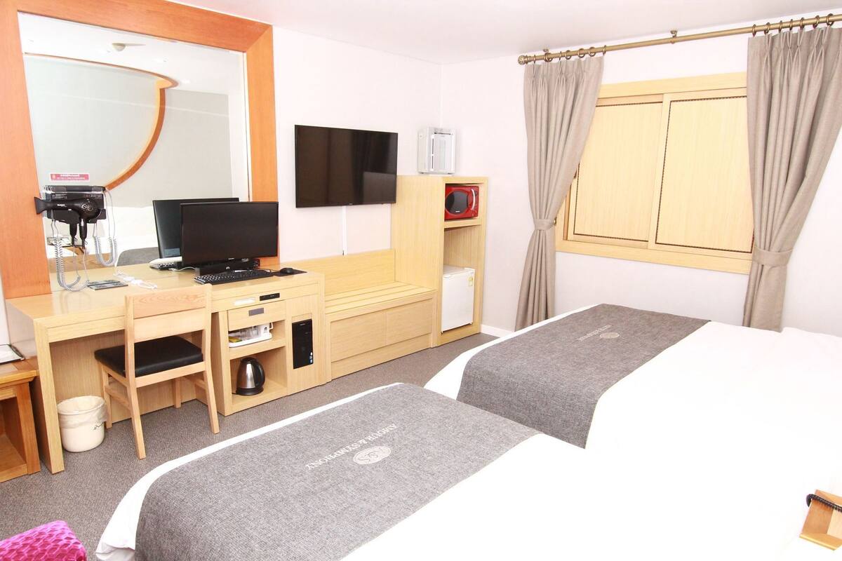 A hotel room with a desk, a television, a minibar, a bed, and a window with curtains.