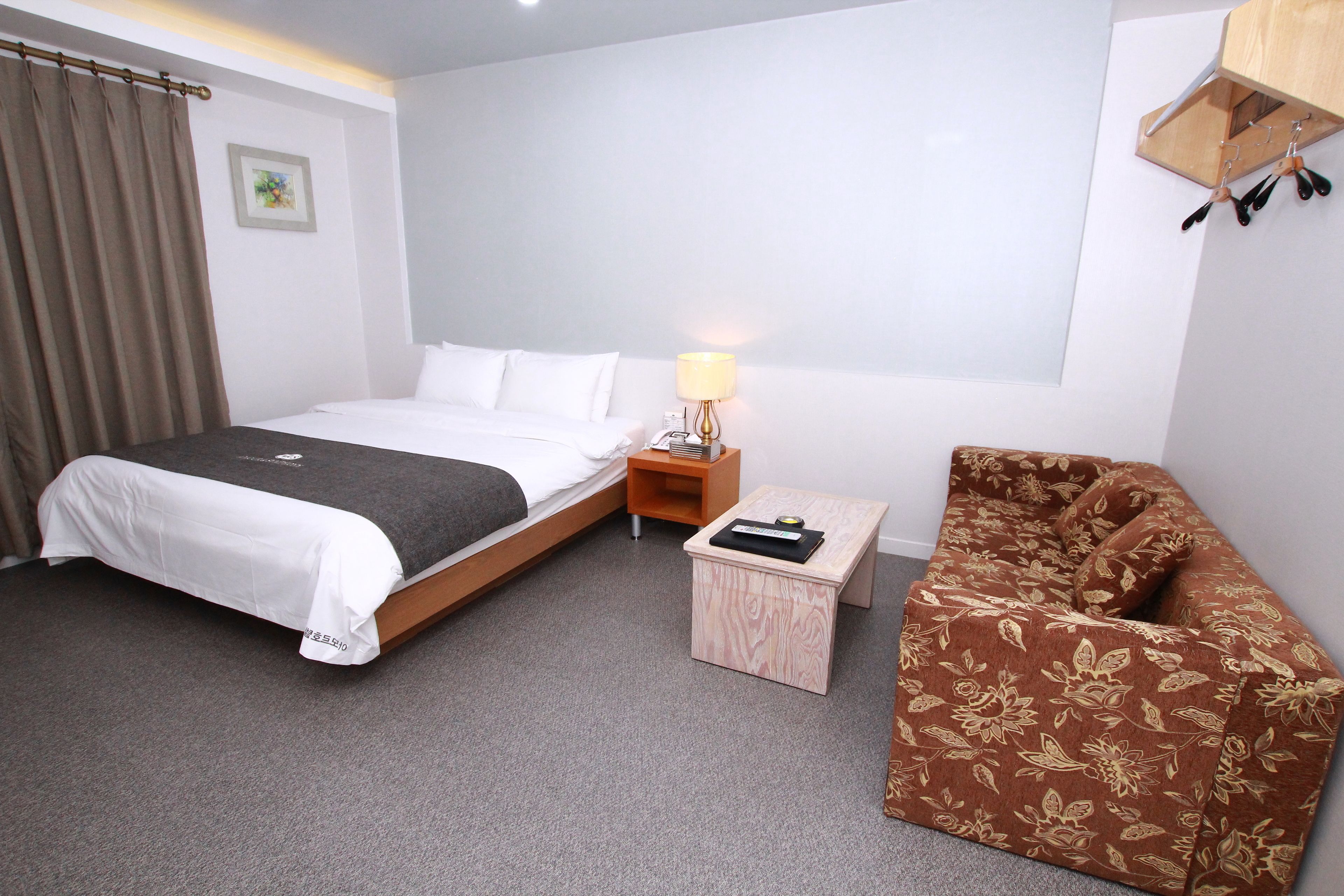 A hotel room with a bed, a nightstand with a lamp, a wooden side table, and a floral-patterned chair.