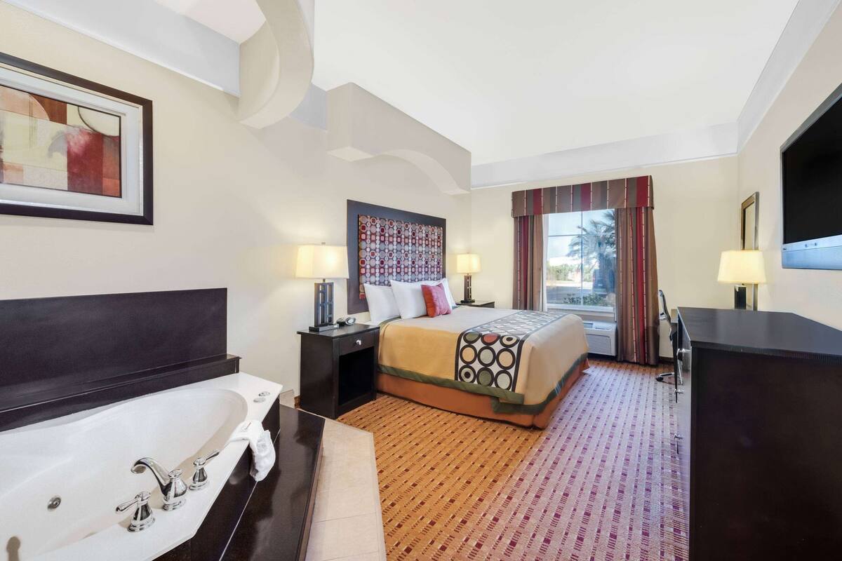 Een hotelkamer met een groot bad, een bed met behang, twee bedlampen en een groot raam met gordijnen.