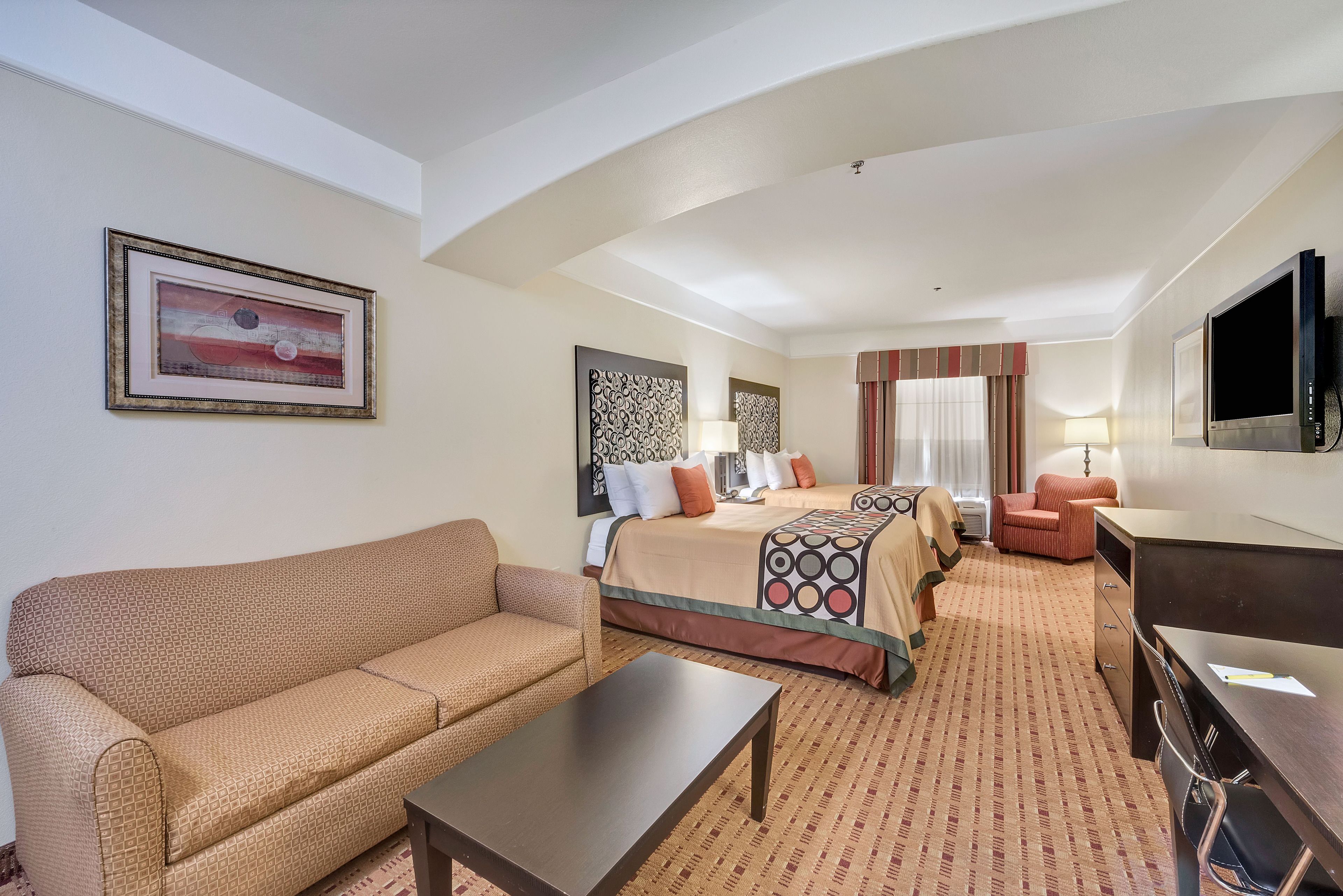 A hotel room with a sofa, a bed with patterned bedding, a desk with a chair, a TV, and artwork on the walls.