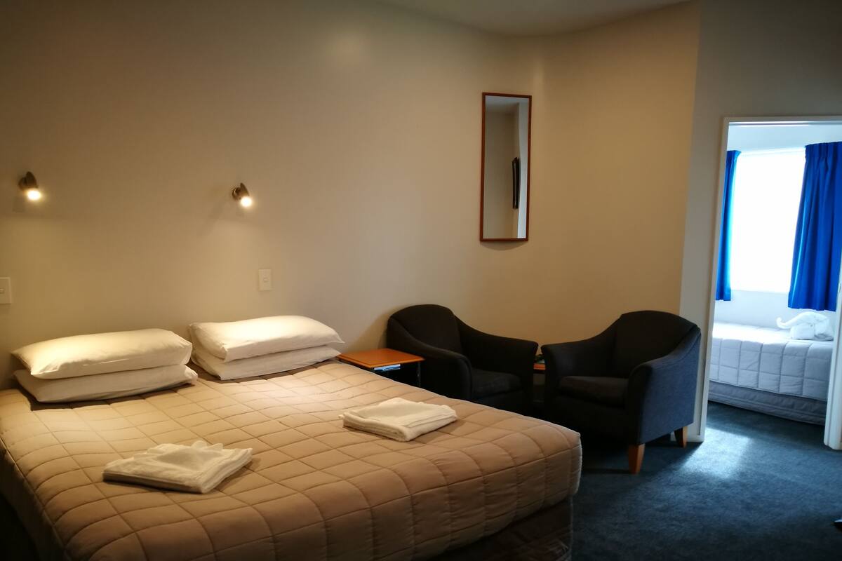 Une chambre d’hôtel avec un grand lit, deux chaises, une petite table, un miroir et une fenêtre avec des rideaux.