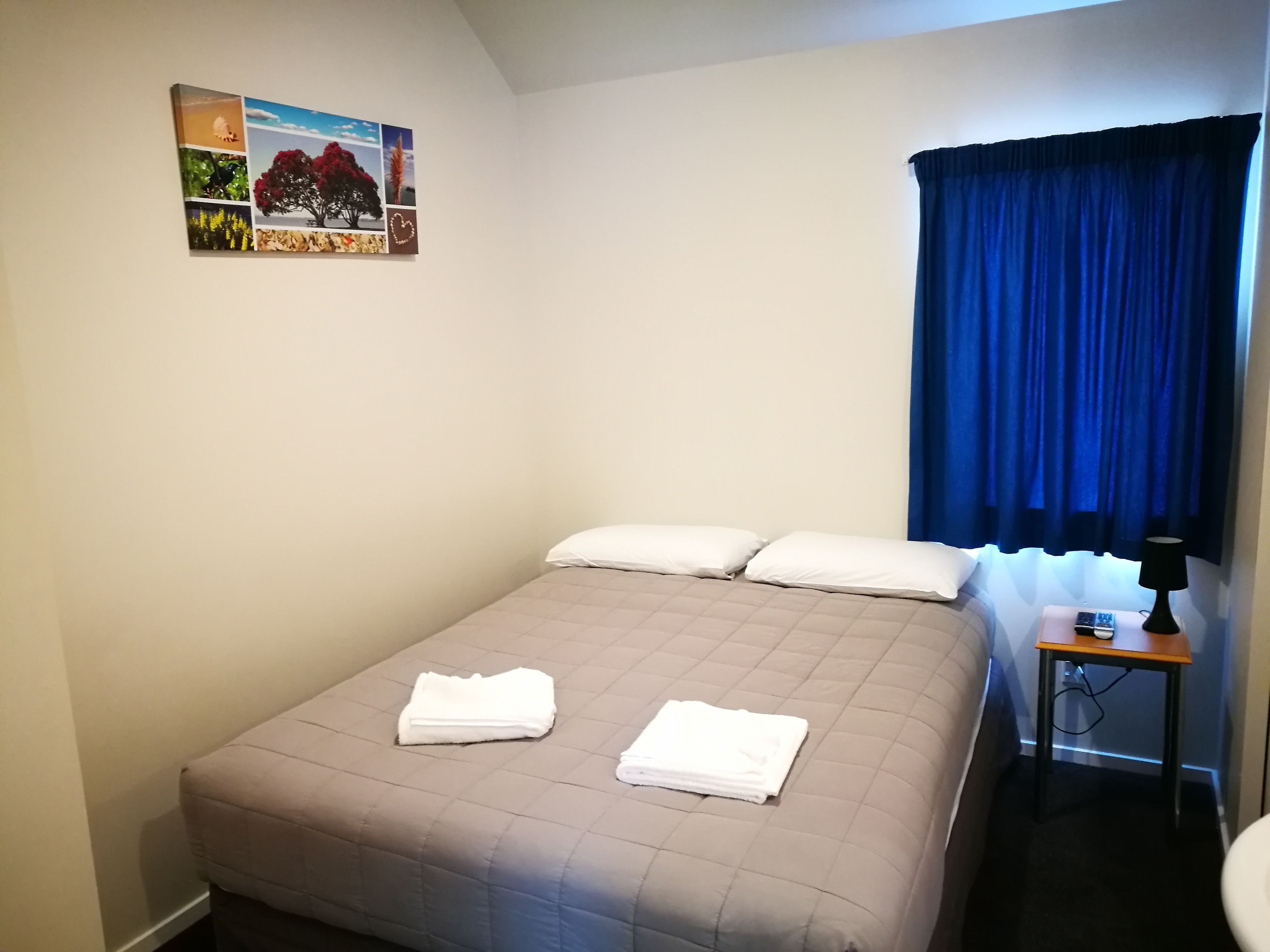 Een netjes opgemaakt bed met twee kussens en gevouwen handdoeken, een klein nachtkastje met een lamp, en een aan de muur hangend schilderij van natuurlandschappen.