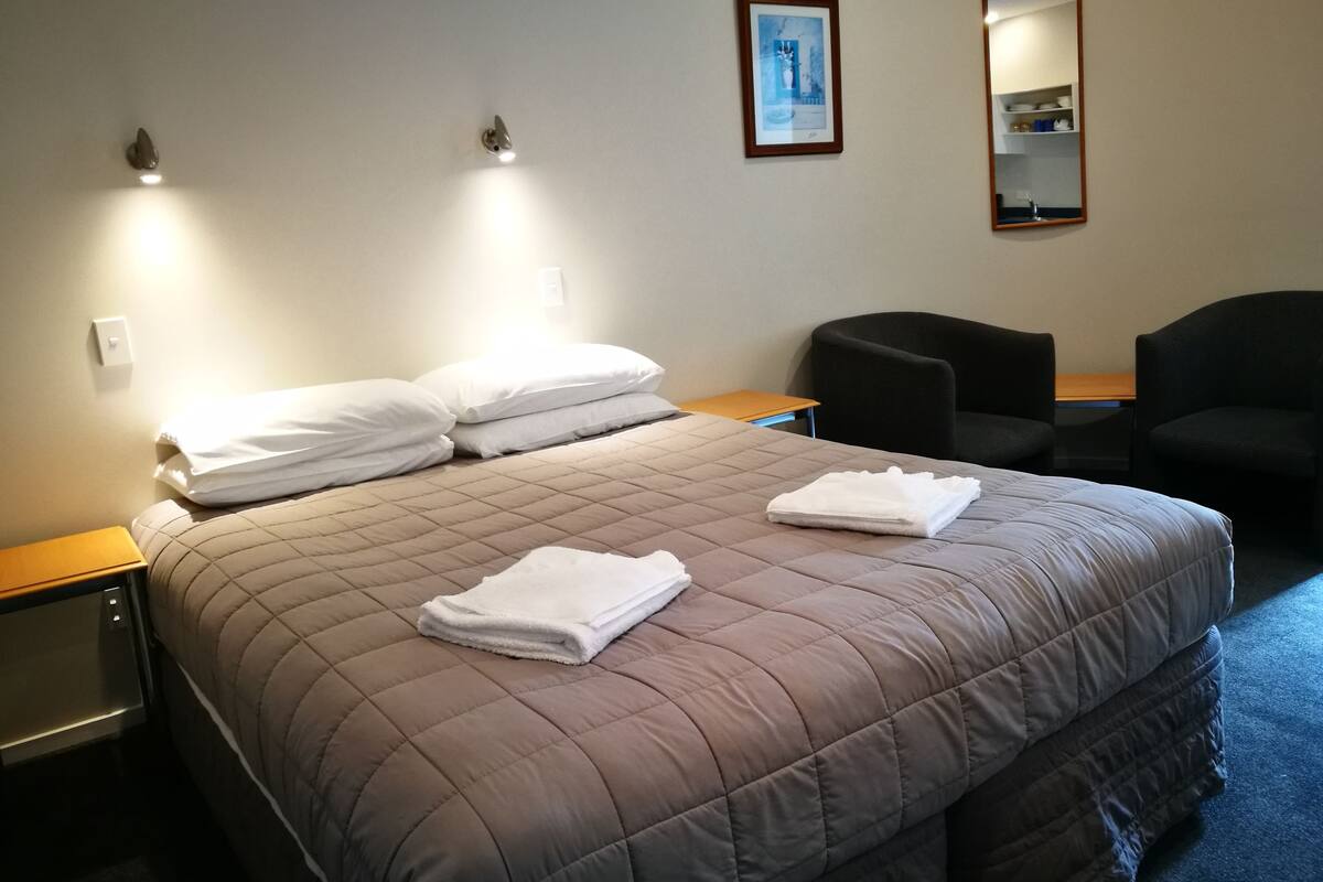 Un lit bien fait, recouvert d’une couverture marron, avec deux oreillers blancs et des serviettes blanches pliées posées sur le lit. On aperçoit une petite table à côté du lit, une chaise noire et un miroir accroché au mur.