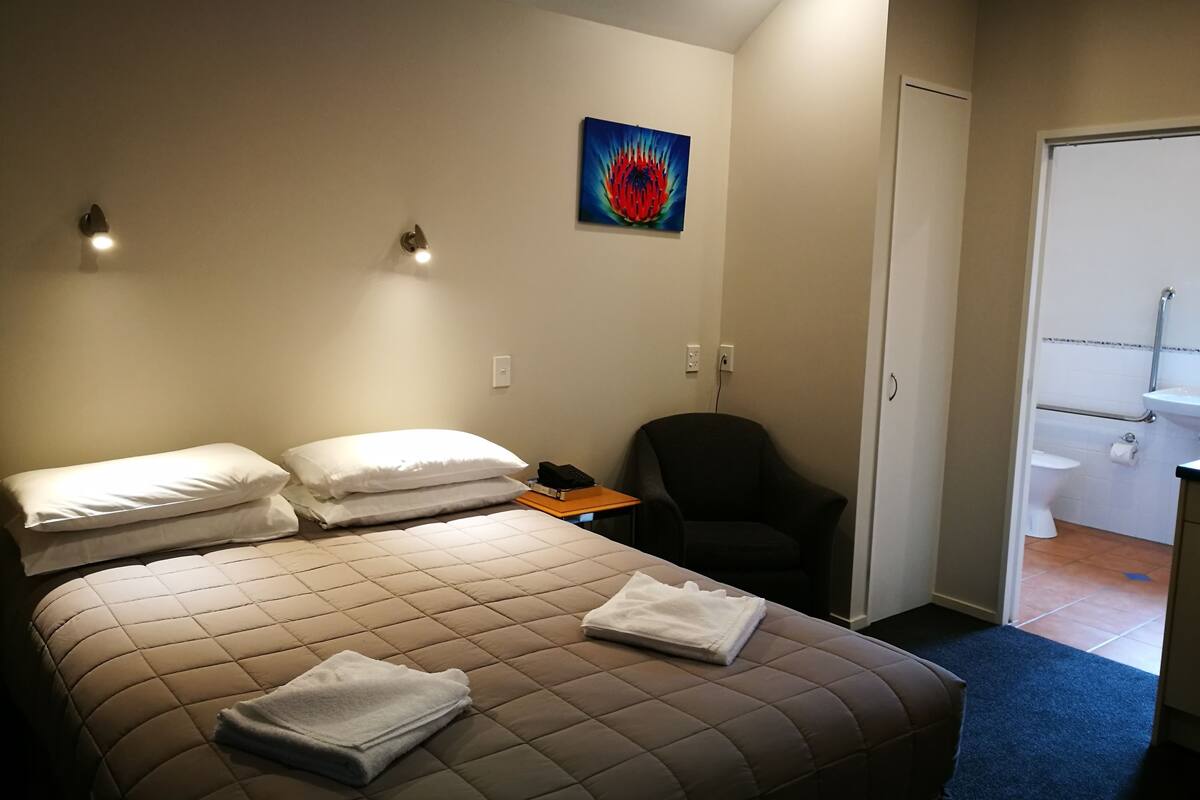 Une chambre d’hôtel avec un lit, des oreillers, une chaise, un tableau et une salle de bain visible à travers une porte ouverte.