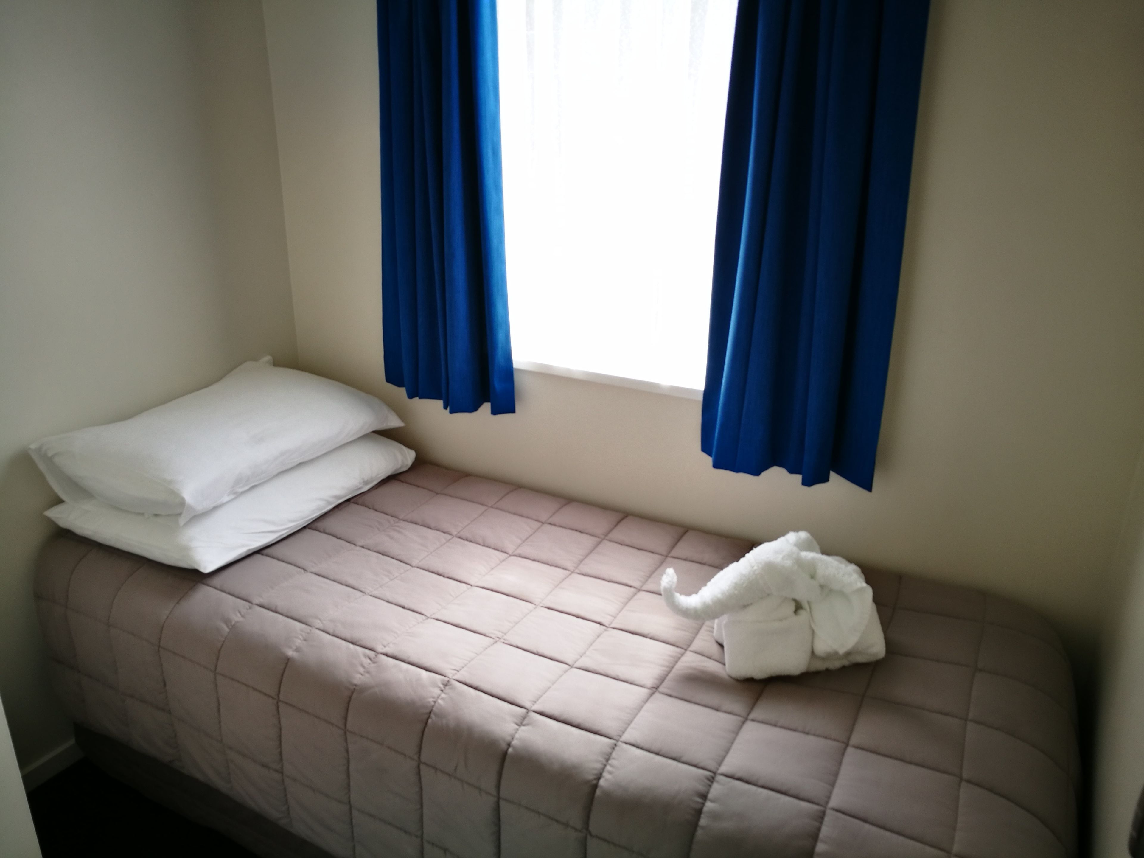 Un lit simple avec un matelas matelassé de couleur beige, des oreillers blancs et une serviette blanche posée sur le côté. Des rideaux bleus sont tirés de part et d’autre d’une fenêtre.