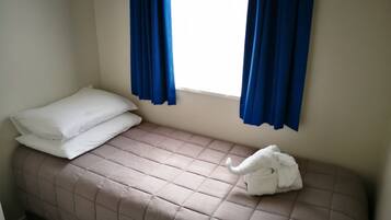 Un lit simple avec un matelas matelassé de couleur beige, des oreillers blancs et une serviette blanche posée sur le côté. Des rideaux bleus sont tirés de part et d’autre d’une fenêtre.