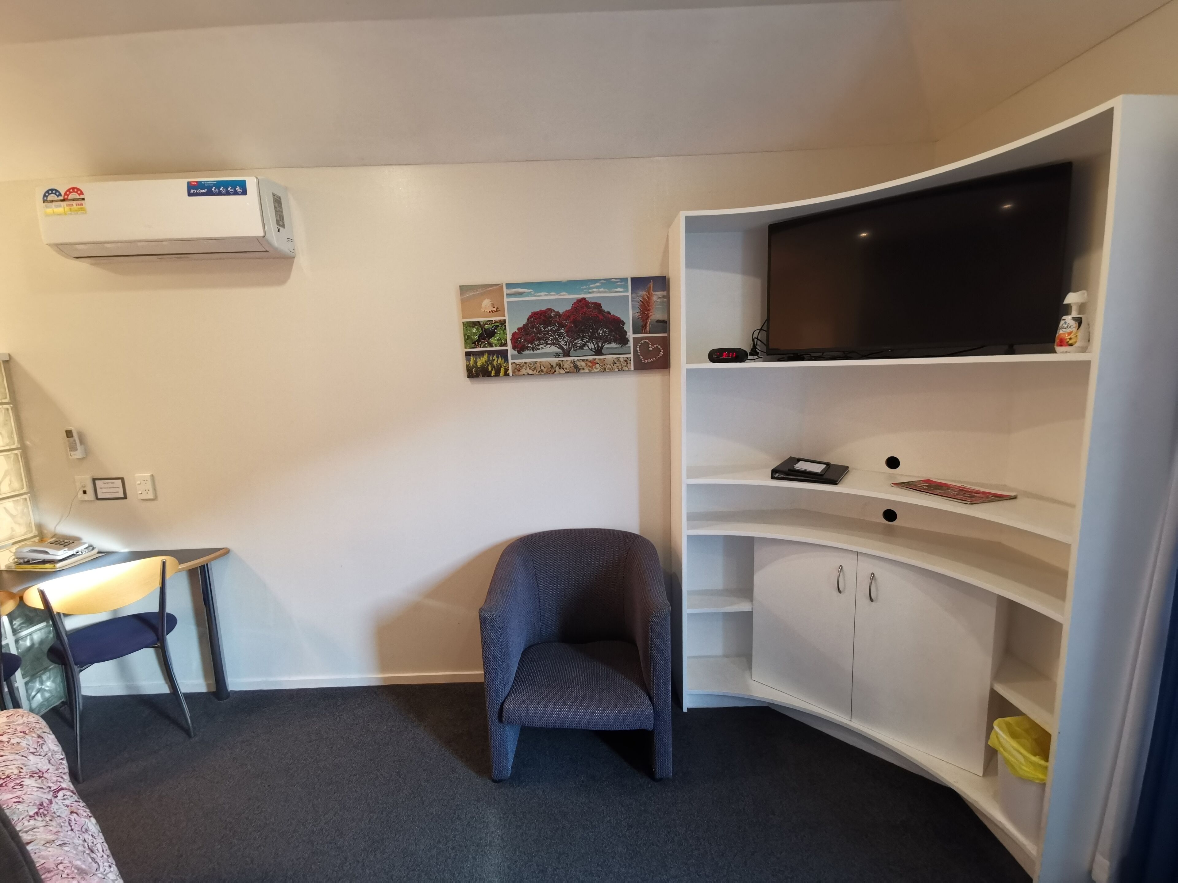 A small, neatly arranged room with a desk, chair, air conditioning unit, wall-mounted TV, and a cabinet with shelves.
