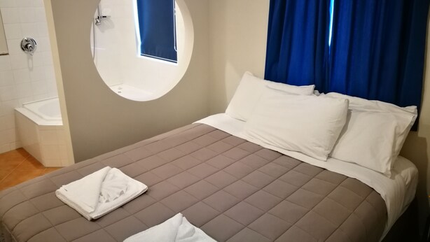 Habitación de hotel con una cama, un espejo redondo, una ventana con cortinas azules y un ventanal pequeño encima del espejo.
