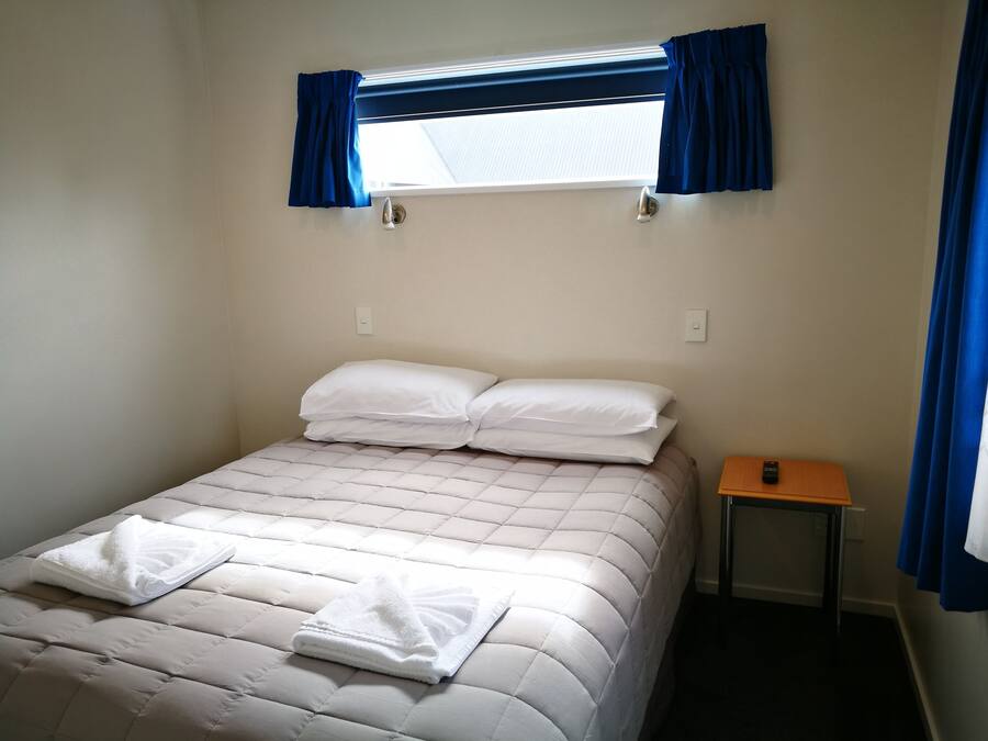 A neatly made bed with white linens and a folded towel, a small bedside table with a remote control, and a window with blue curtains.