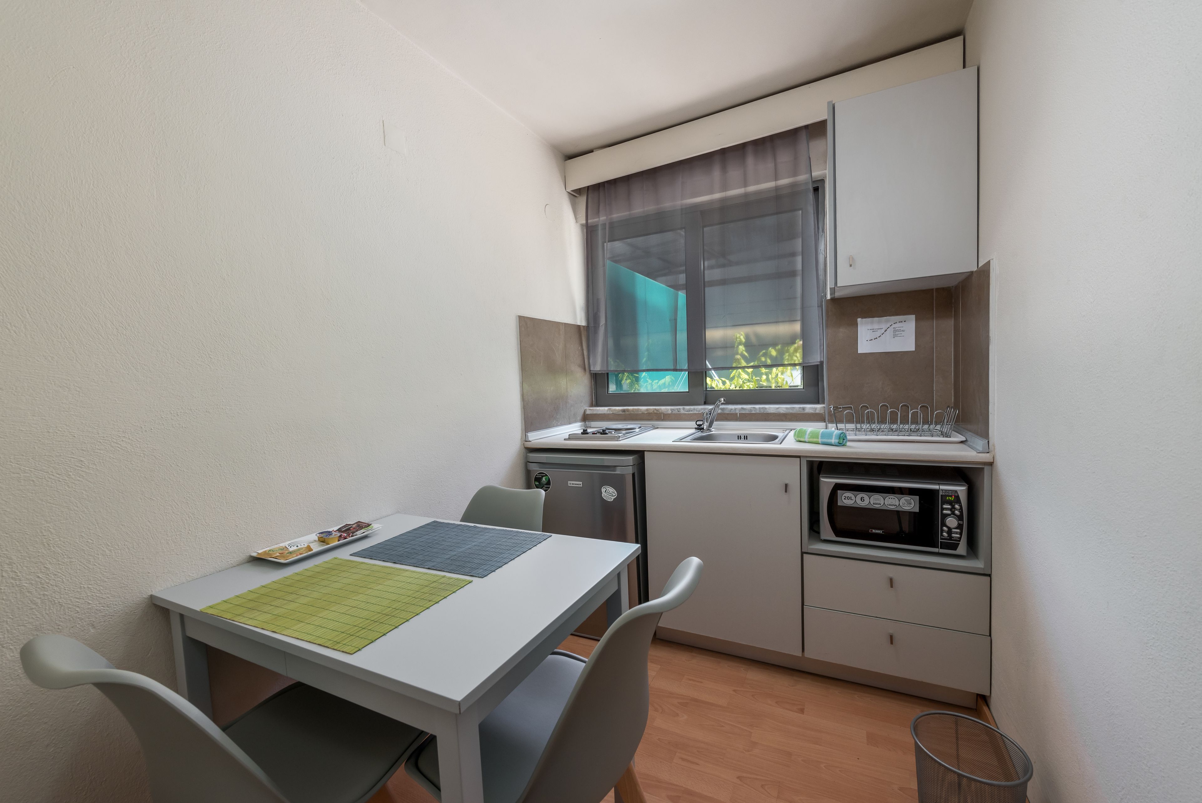 A compact kitchenette with a microwave, sink, and small refrigerator. There is a dining table with chairs and a window with curtains.