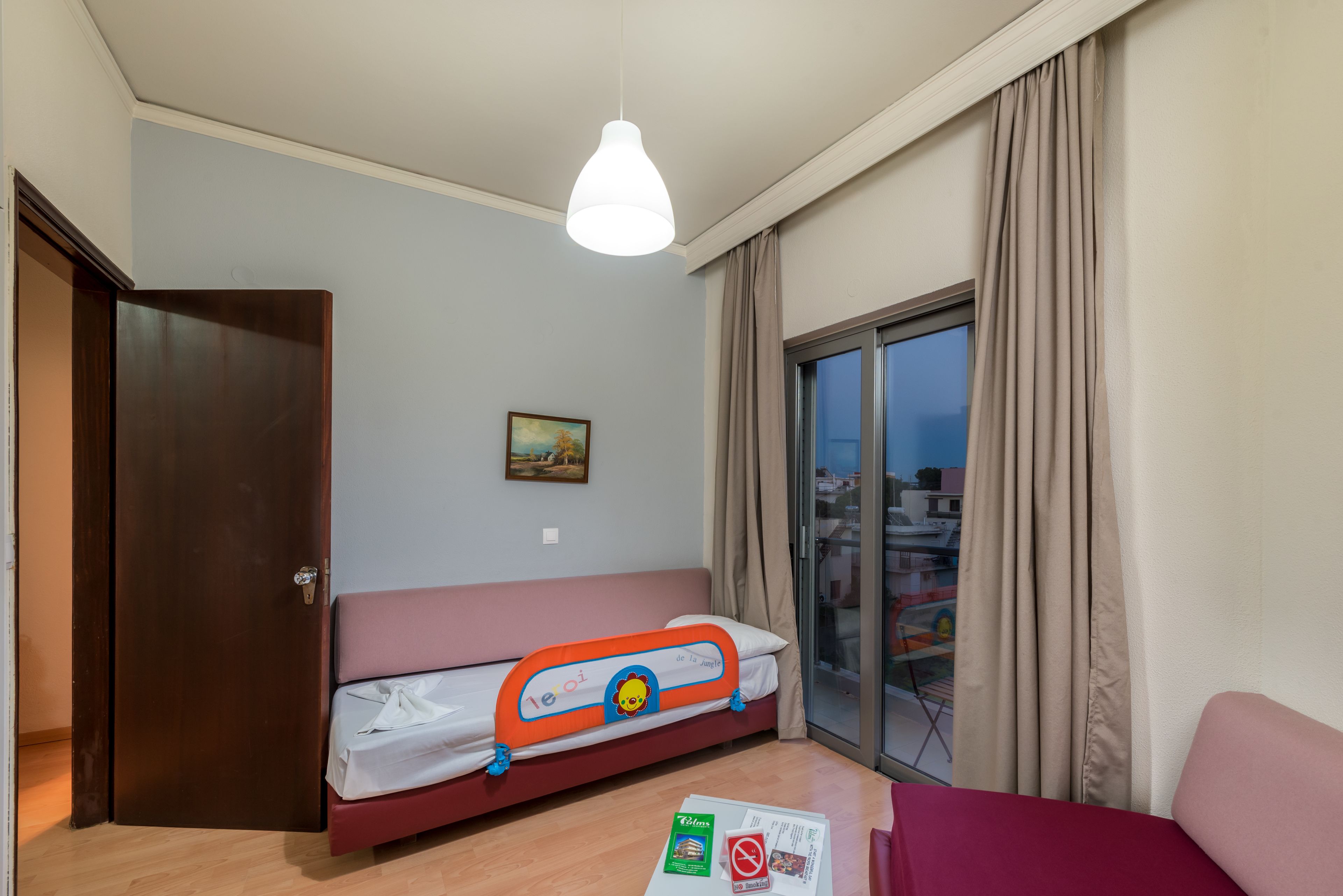 Um quarto com cama, janela com cortinas, porta e mesa com uma revista.