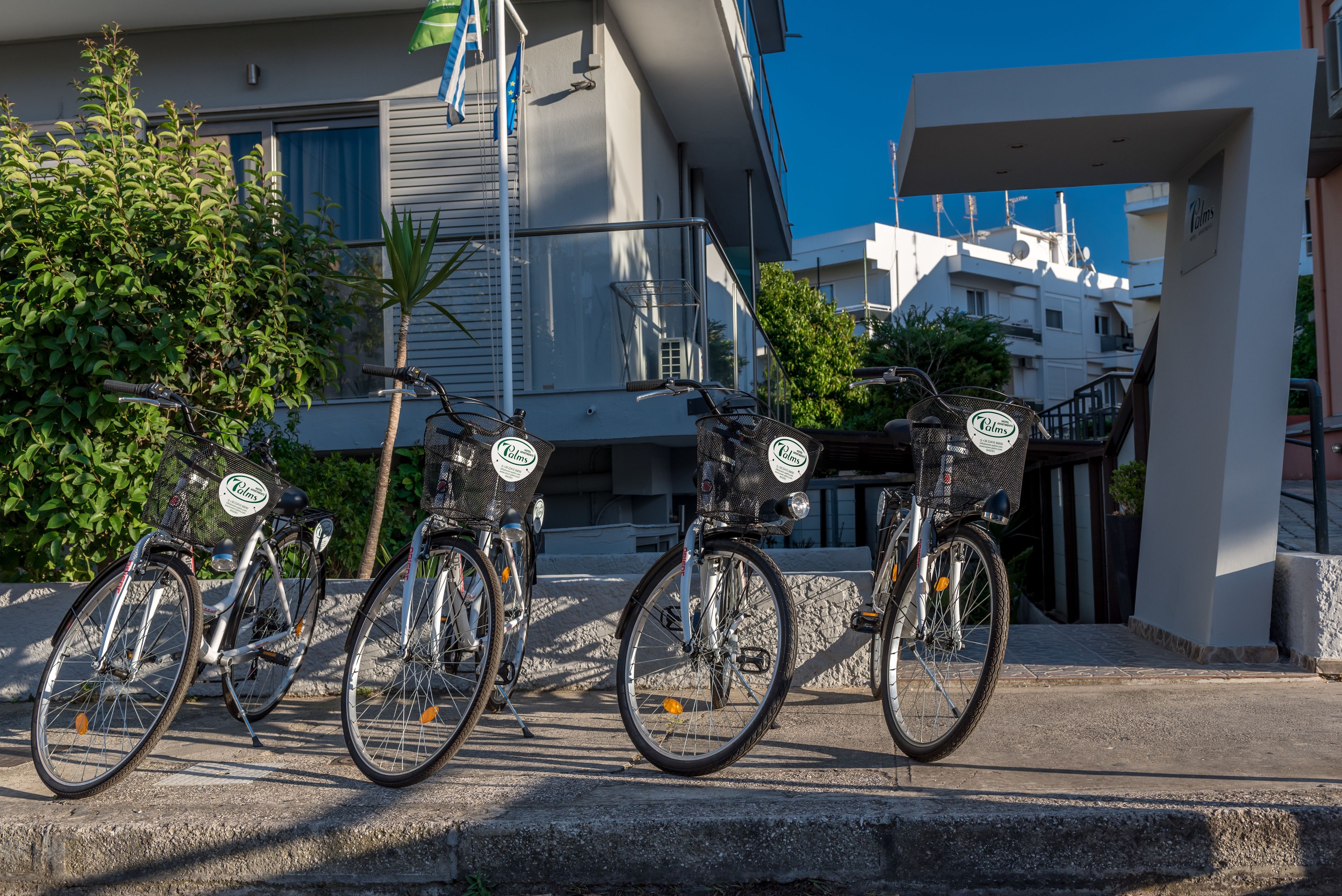 A row of bicycles with baskets, parked on a concrete surface in front of a building with balconies and a clear blue sky in the background.