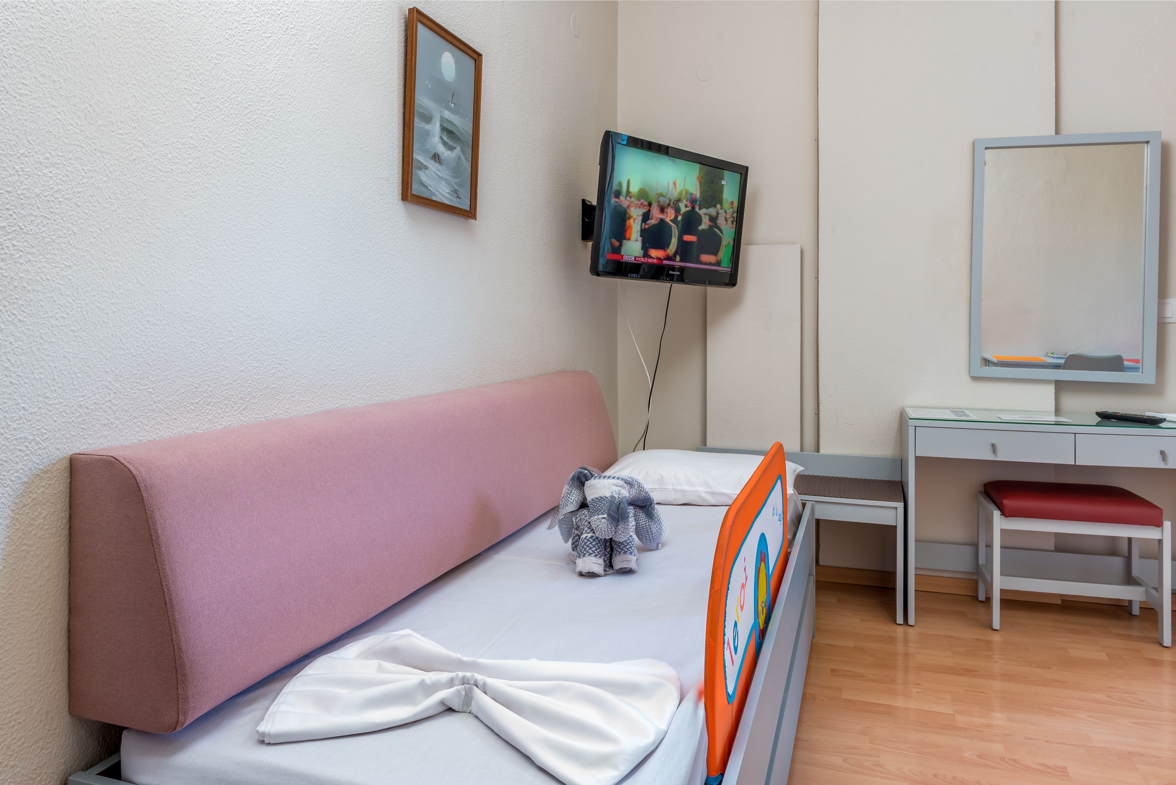 Een kinderkamer met een bed, een bureau, een stoel, een televisie en een ingelijst beeld aan de muur.