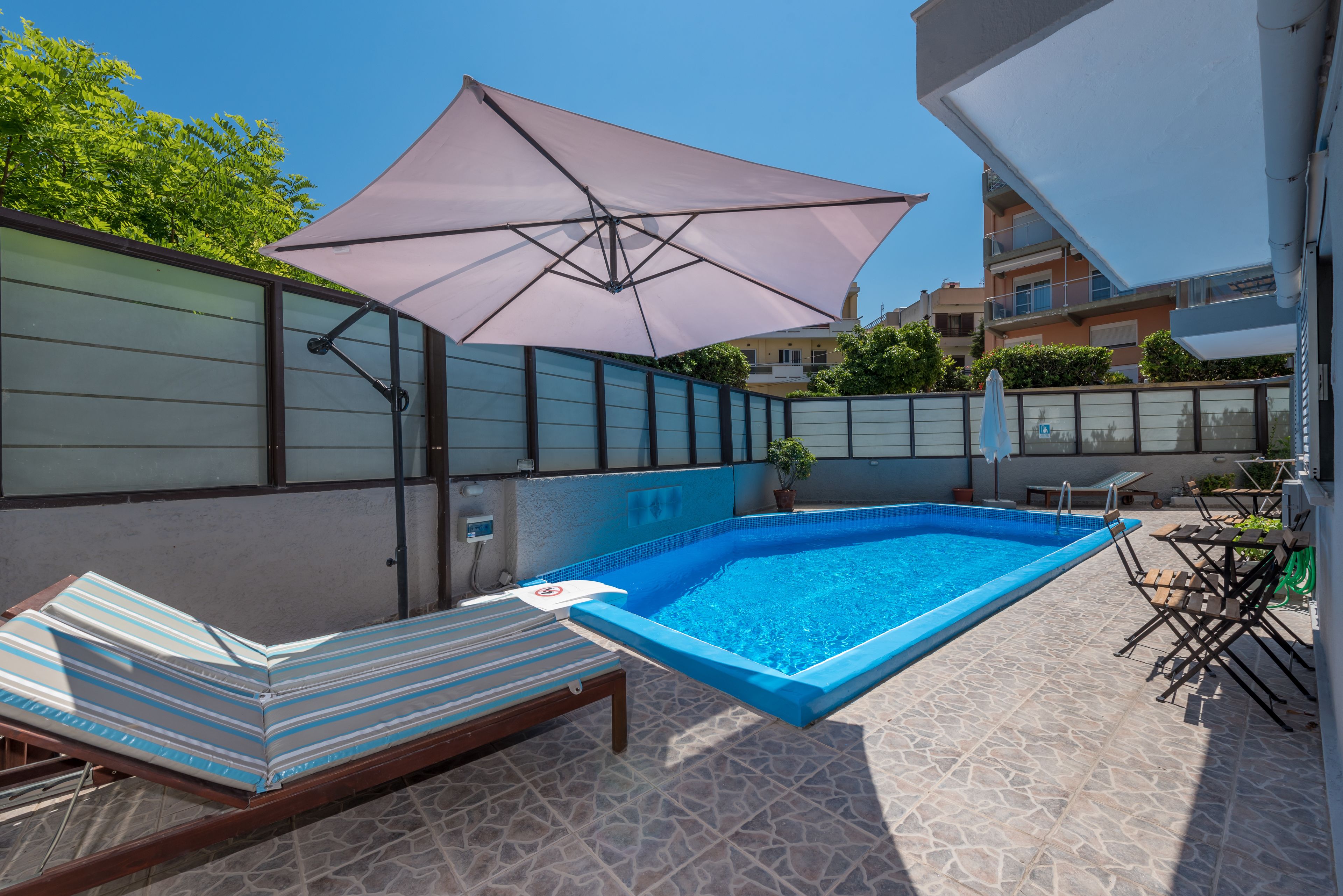 Ein Swimmingpool mit Liegestuhl und Sonnenschirm, umgeben von einer gefliesten Terrasse und einem Zaun.