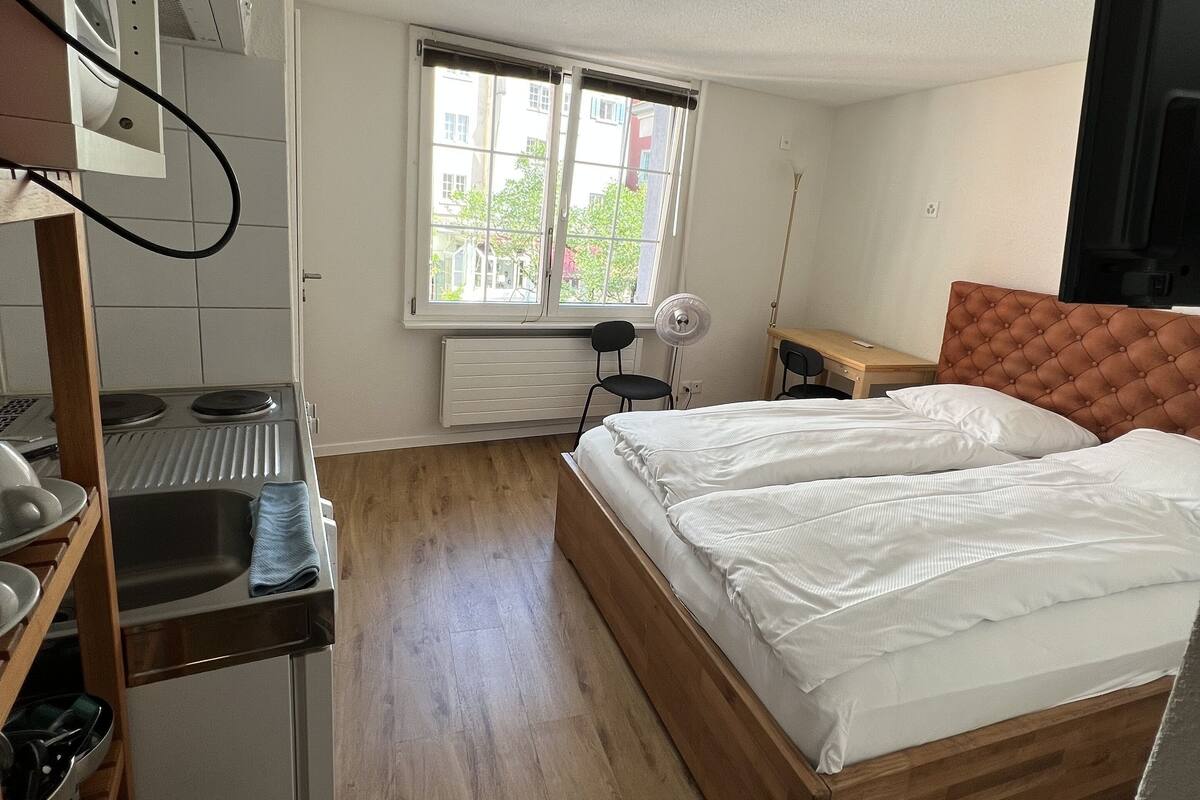 A compact hotel room with a bed, a small desk, a window with a view of buildings, and a radiator.