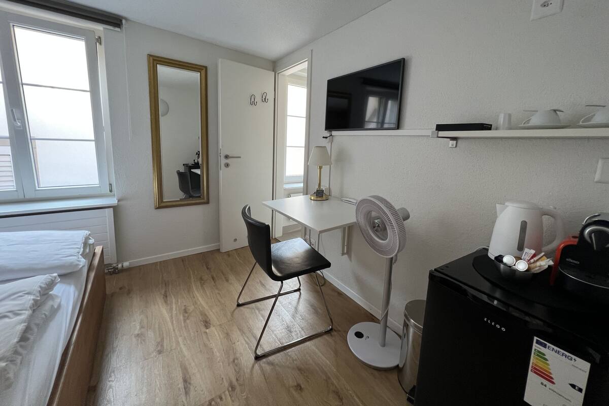 A small, clean room with a bed, a desk, a chair, a TV, a fan, and a coffee maker.