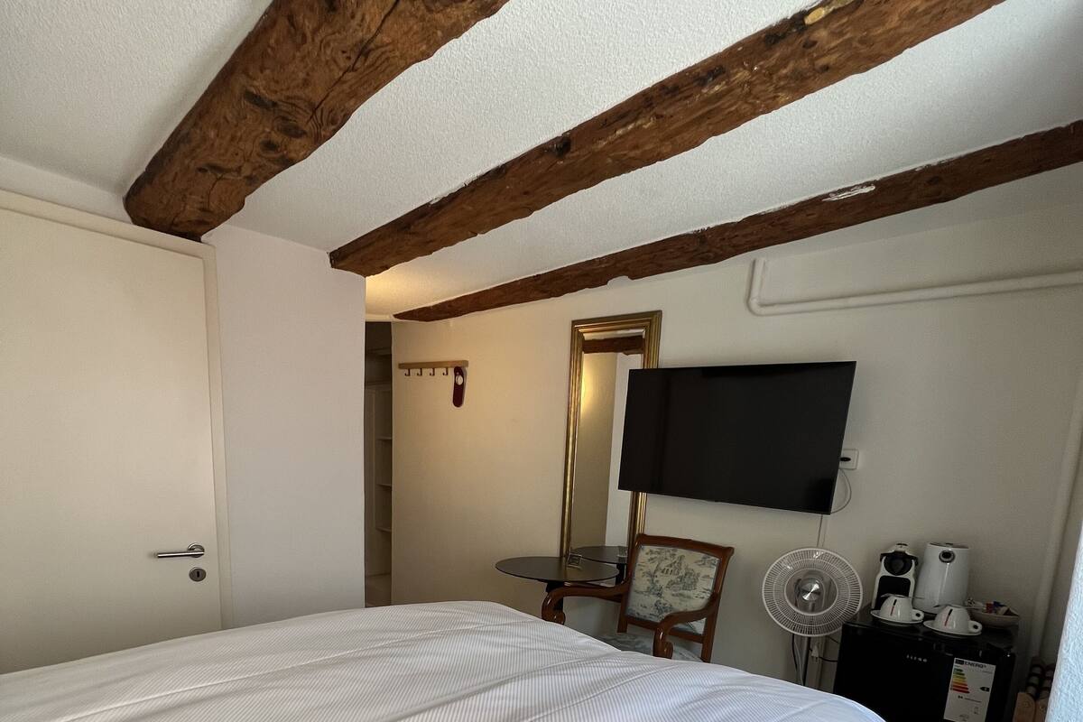 A bedroom with a large bed, wooden beams, a TV, a small table, a chair, a fan, and a refrigerator.