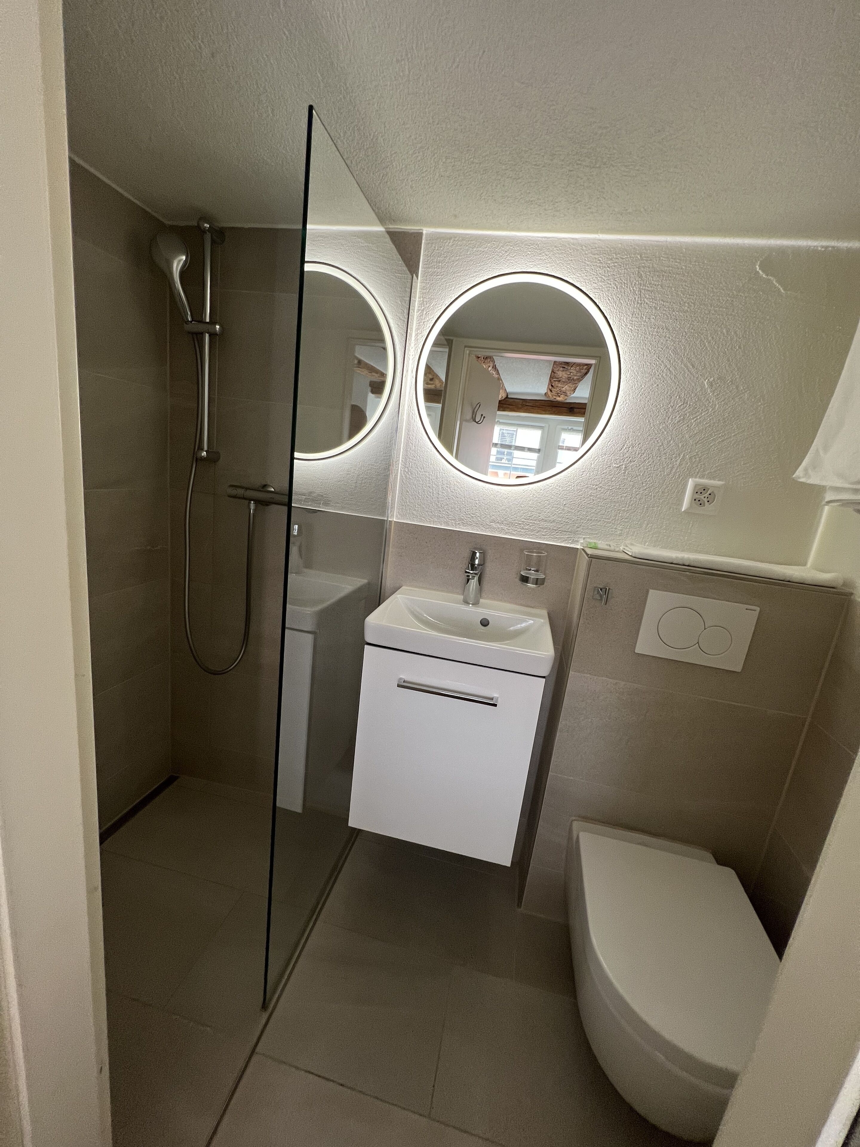 Un baño con cabina de ducha de vidrio, lavamanos, inodoro y dos espejos redondos.
