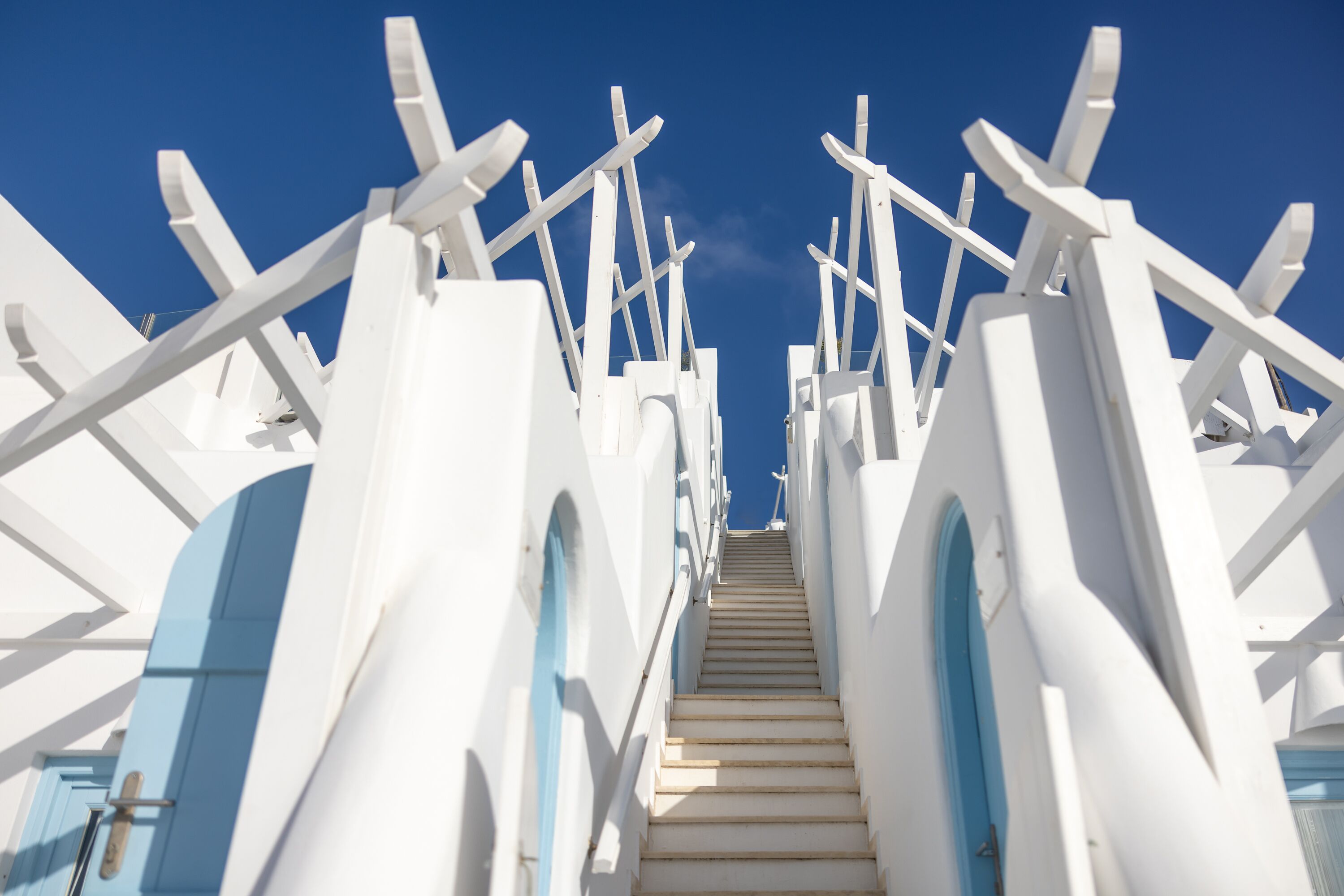 Una escalera que sube con barandillas blancas y puertas azules, rodeada de paredes blancas.