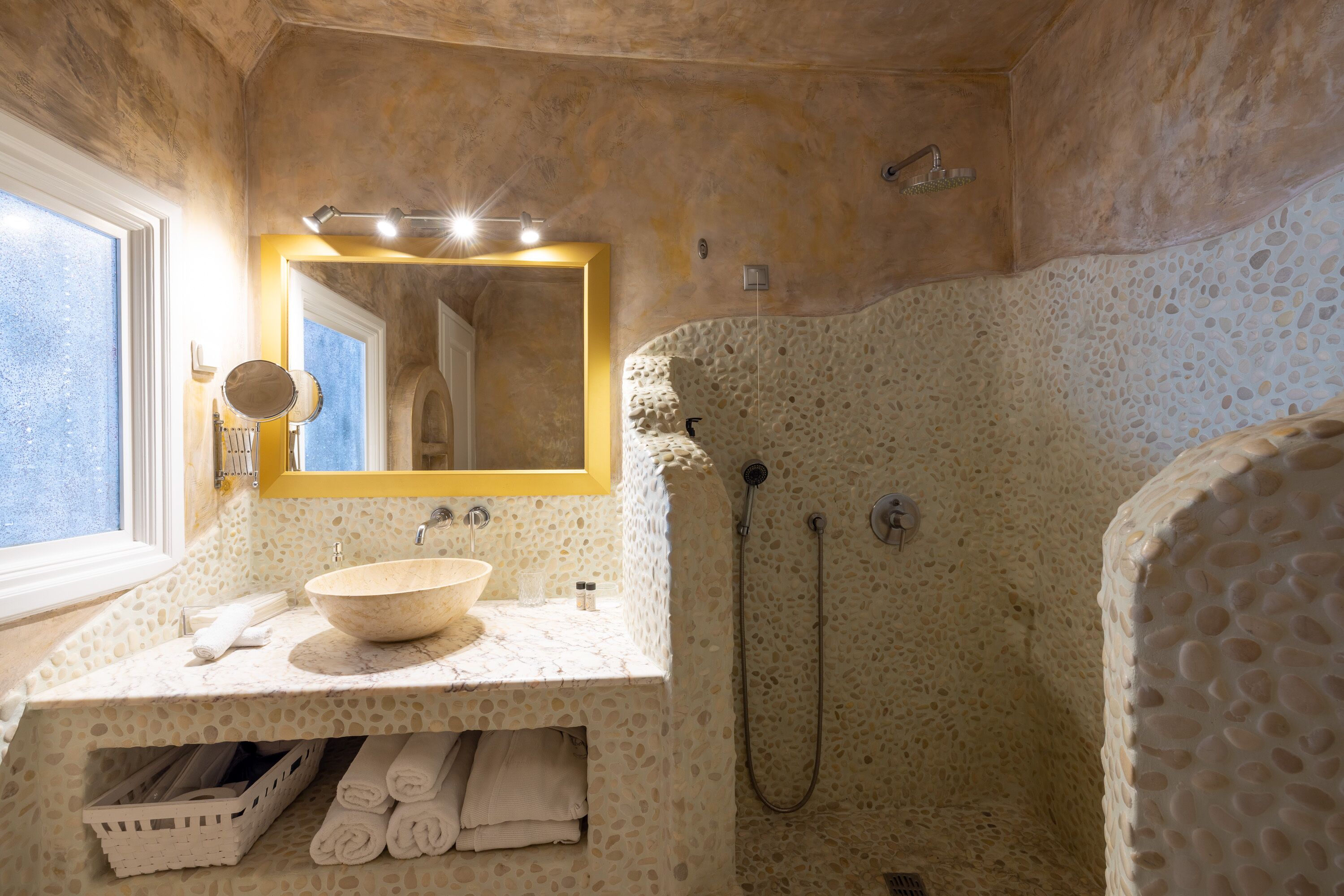 Un baño con pared de textura de piedra, un lavabo, un espejo con marco amarillo y una zona de ducha.