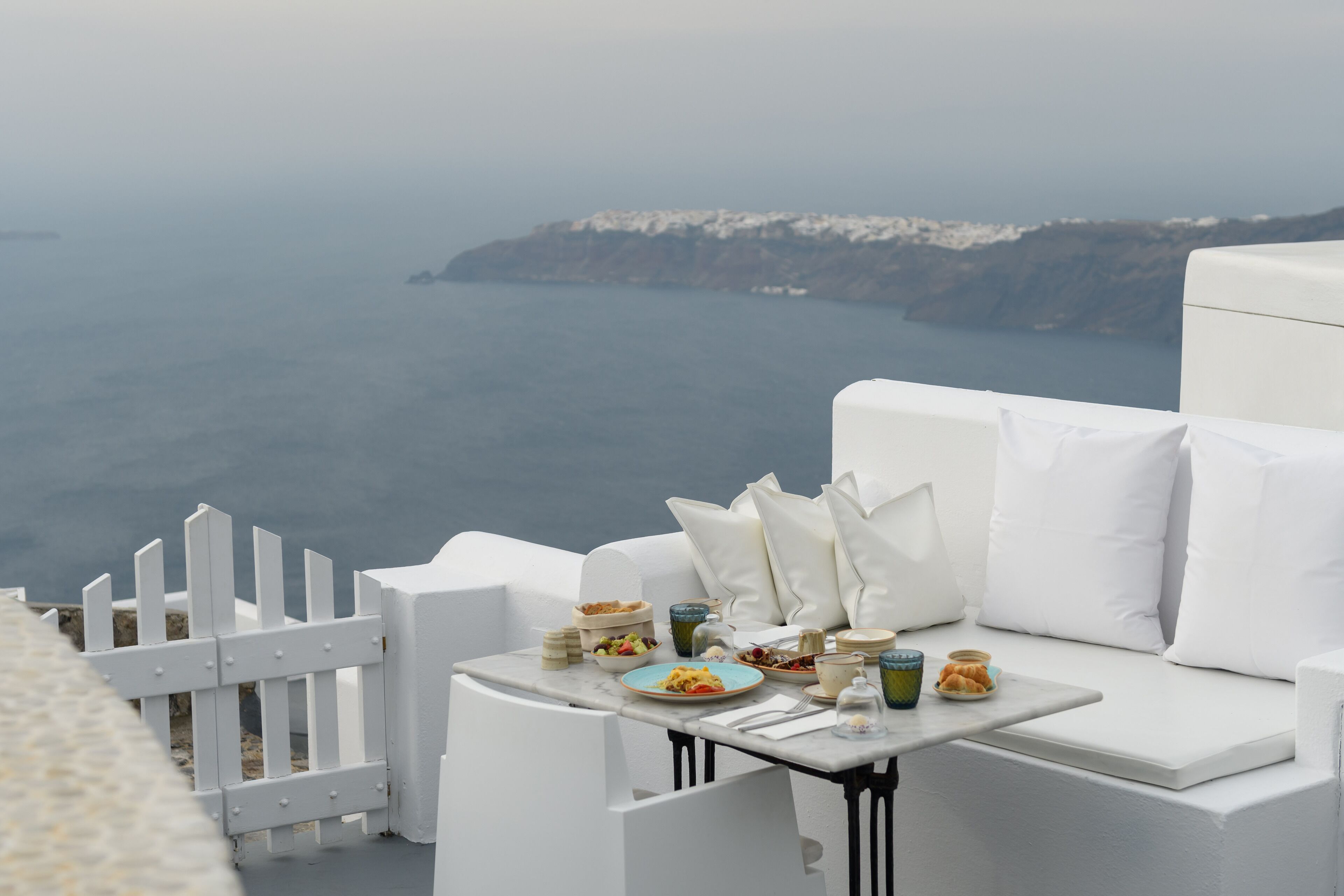 A white outdoor seating area with a table set for breakfast, overlooking a coastal view.