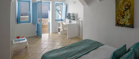A bedroom with a bed, a desk, a chair, and a television. There is a balcony with a view of the sea.