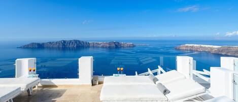 A seaside lounge with white lounge chairs and a clear view of the ocean and a rocky island in the distance.