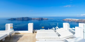 A seaside lounge with white lounge chairs and a clear view of the ocean and a rocky island in the distance.