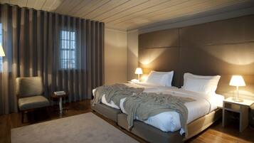Quarto de hotel moderno com uma cama grande, duas mesas de cabeceira com luminárias, uma cadeira e vista para o exterior através de uma janela com cortinas.