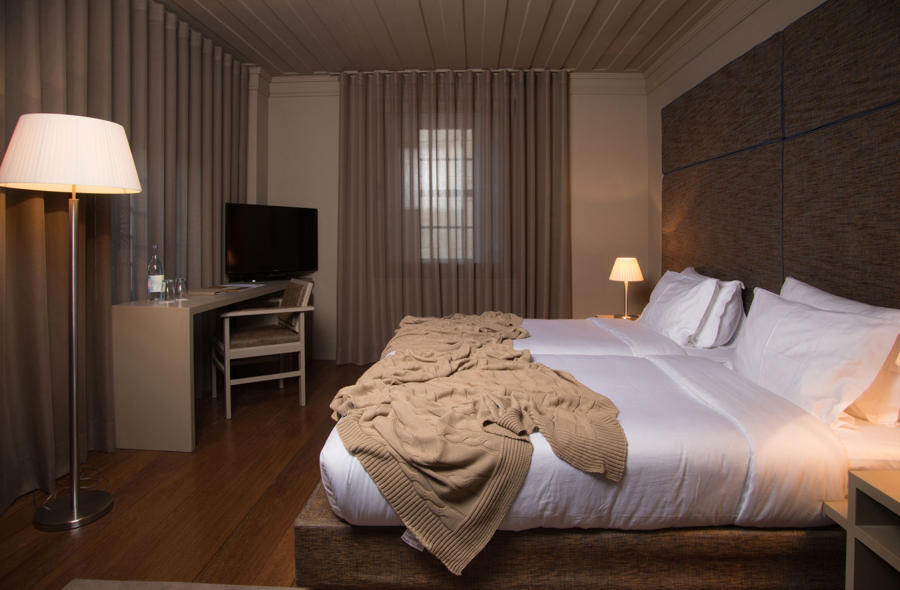 Une chambre d’hôtel avec un lit, un bureau avec une télévision, une lampe et une fenêtre avec des rideaux.