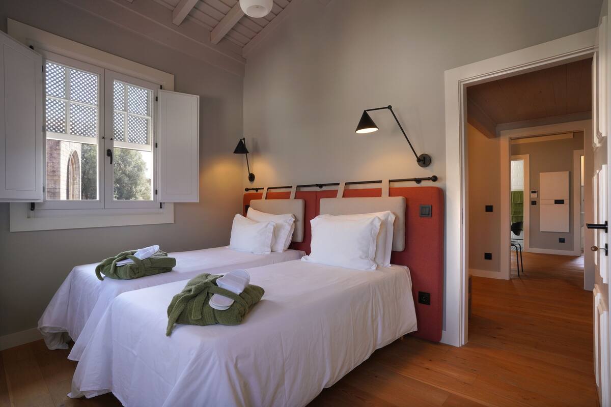 A hotel room with two beds, a window with a lattice design, and a hallway leading to another room.