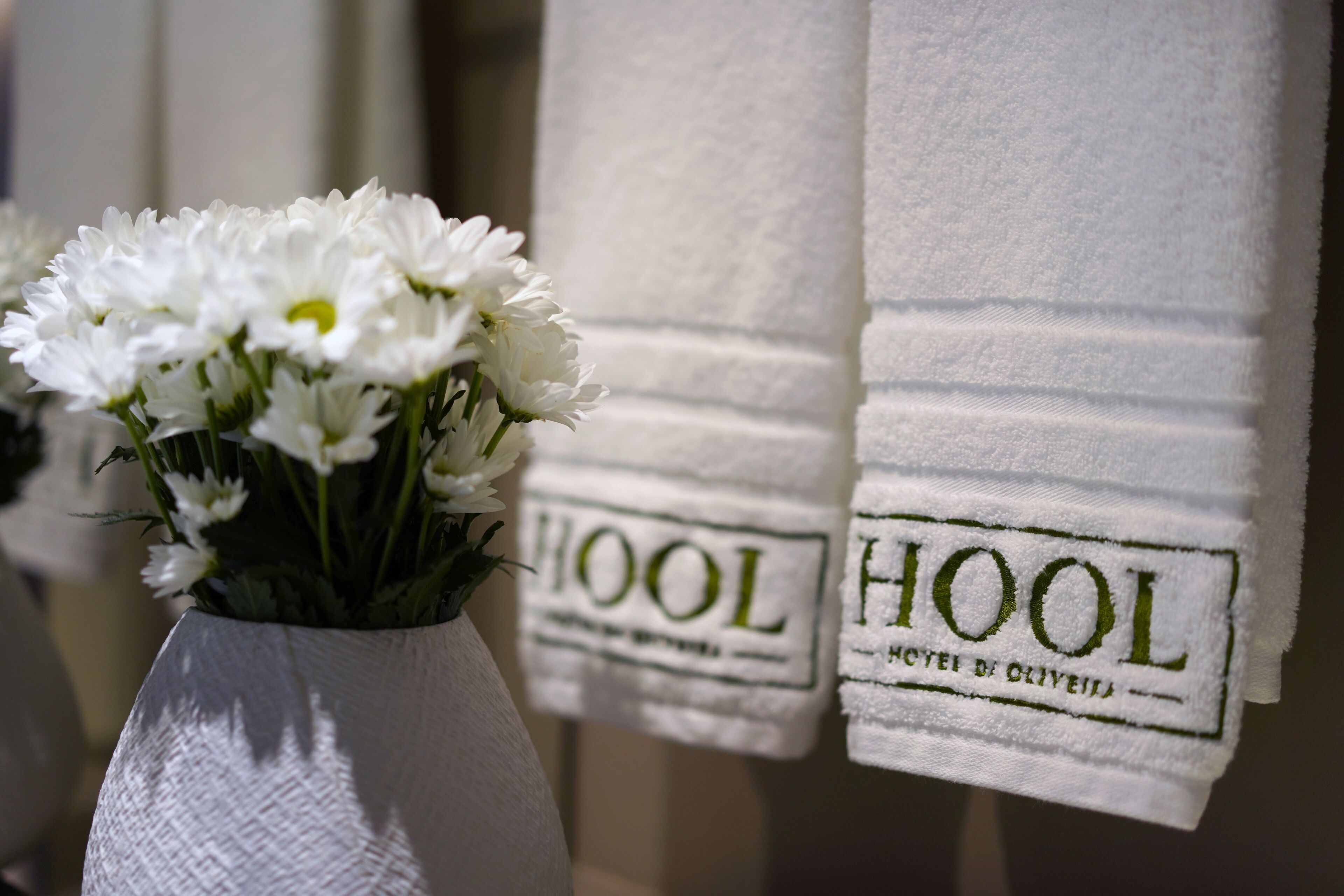 Un jarrón blanco con flores blancas y dos toallas blancas con el bordado 'HOOL'.