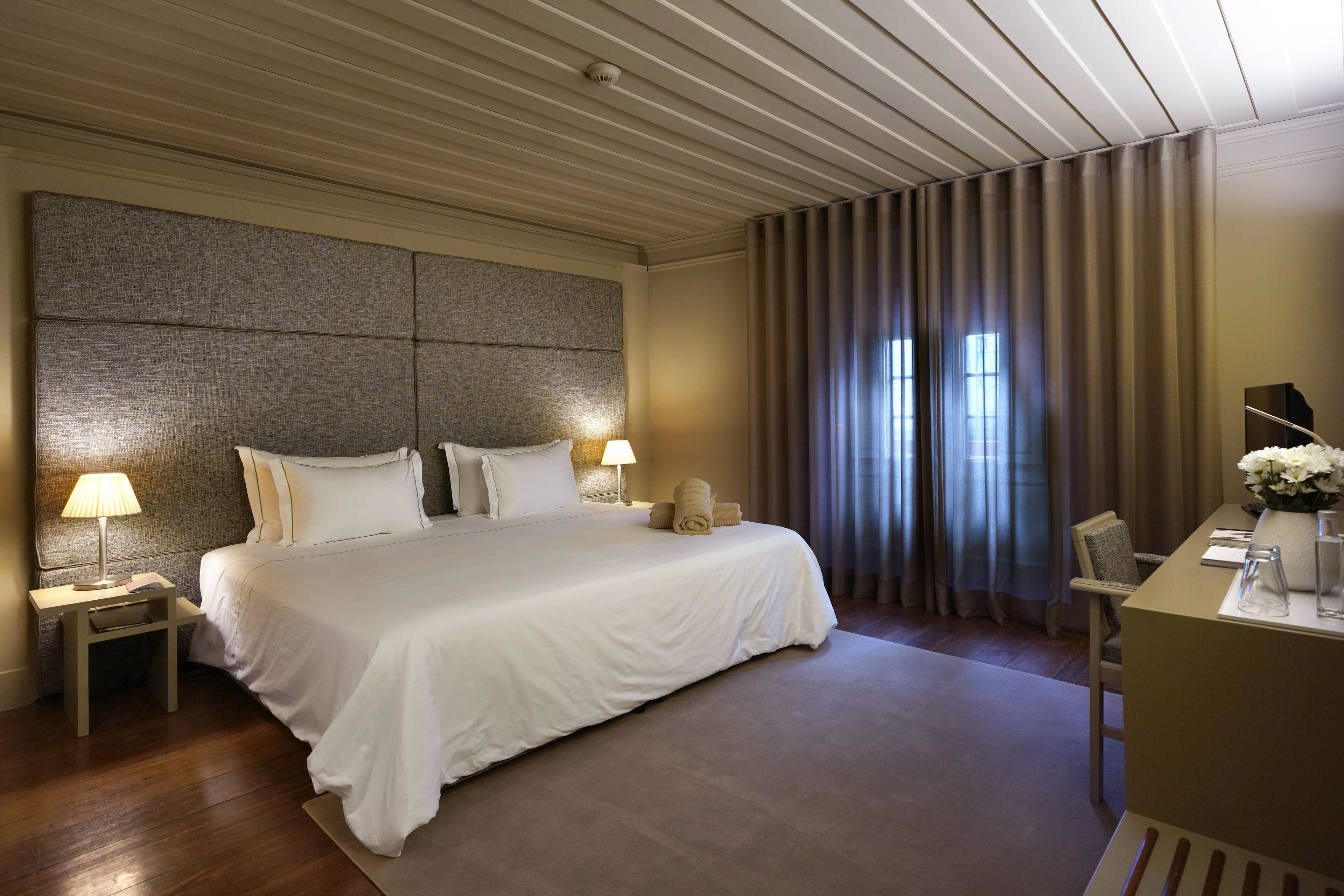 Habitación de hotel con una cama grande, dos mesitas de noche con lámparas, un escritorio con silla y una ventana con cortinas.